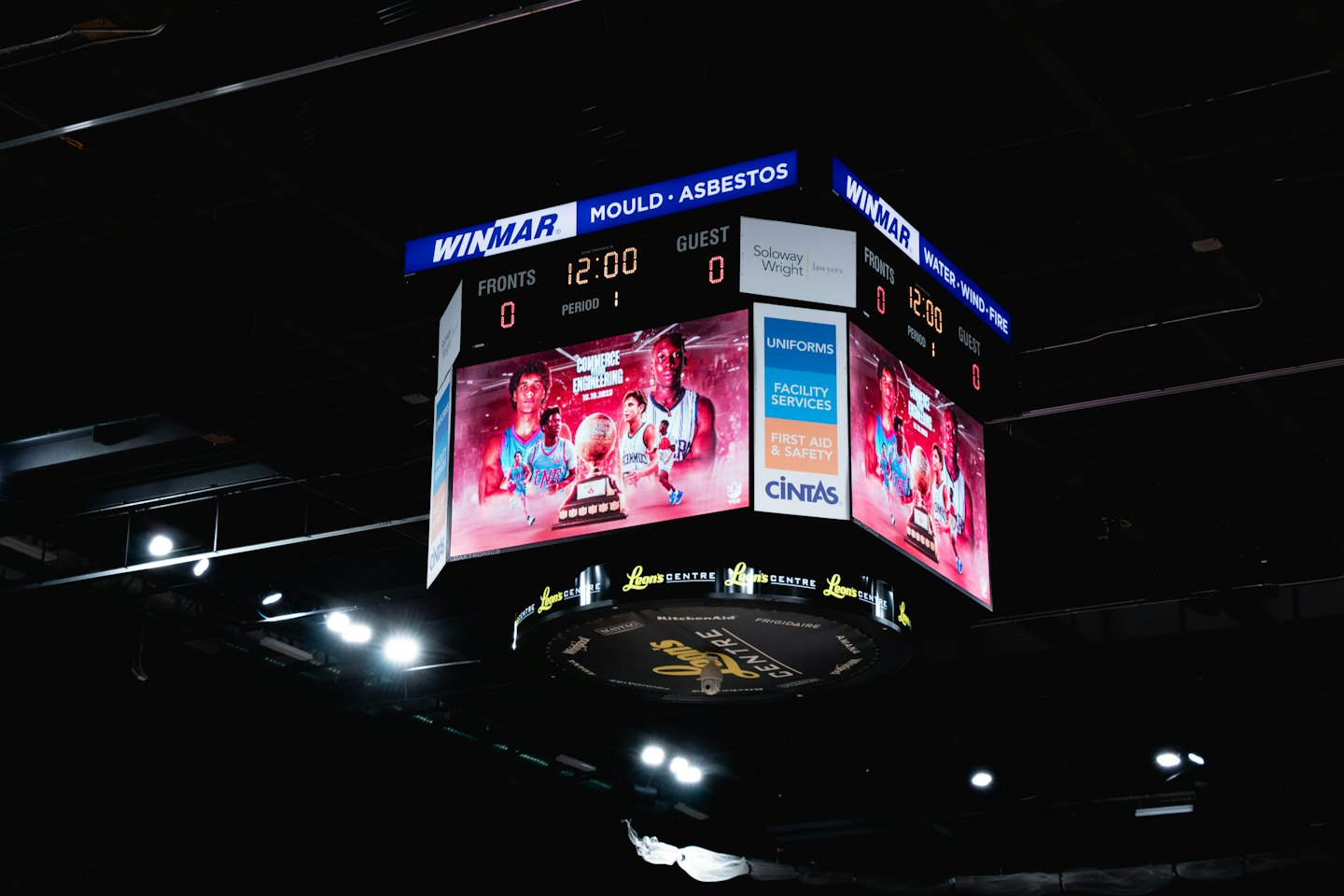 A large multi-sided LED scoreboard hangs in a dark arena.
